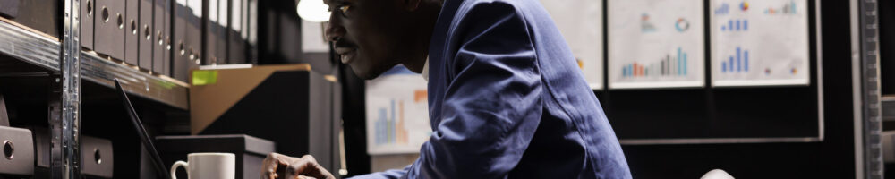 Secretary looking at bookkeeping files African american secretary looking at bookkeeping files, analyzing administrative documents late at night in storage room. Businessman discovering bureaucracy report, checking accountancy paperwork