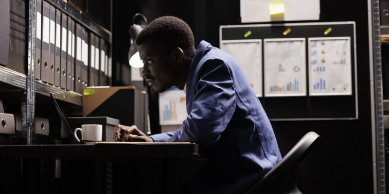 African american secretary looking at bookkeeping files, analyzing administrative documents late at night in storage room. Businessman discovering bureaucracy report, checking accountancy paperwork