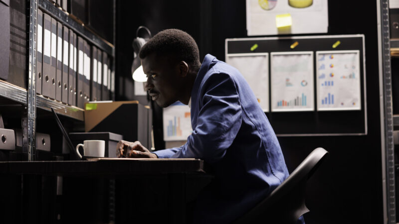 African american secretary looking at bookkeeping files, analyzing administrative documents late at night in storage room. Businessman discovering bureaucracy report, checking accountancy paperwork