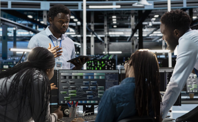 Engineers doing brainstorming, programming in server farm housing infrastructure storing datasets. Teamworking colleagues run code on PC to repair data center racks doing computational operations